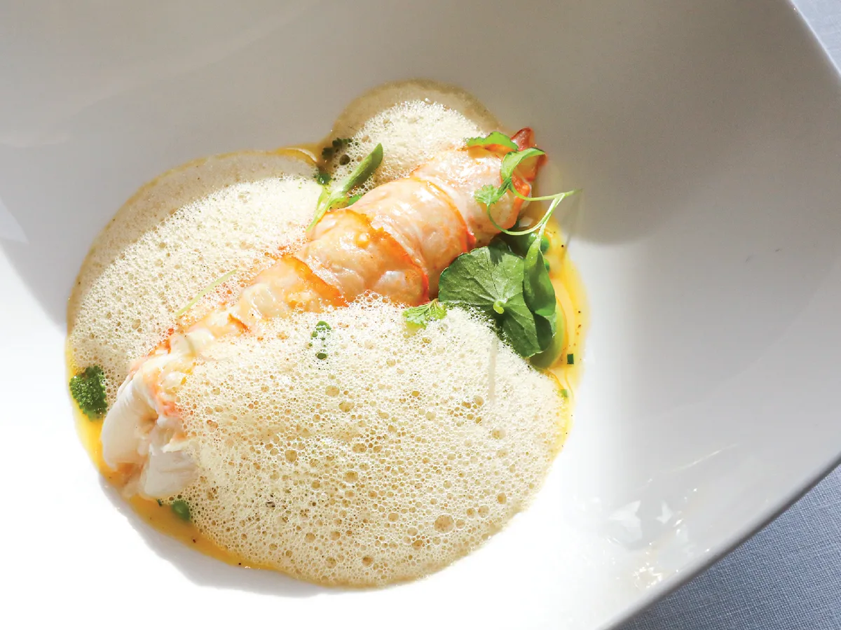 กุ้งลังกูสตีนคลุมฟองซอสบางเบา ตกแต่งด้วยใบสมุนไพรในจานสีขาว