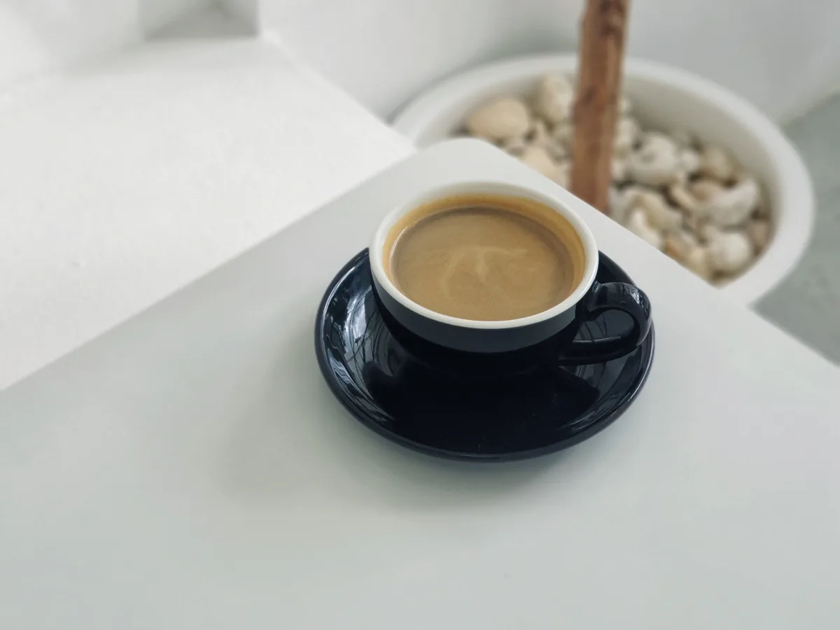 กาแฟร้อนในถ้วยสีน้ำเงินบนโต๊ะขาวข้างกระถางกิ่งไม้