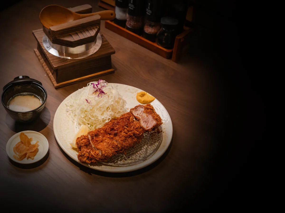 ชุดหมูทอดทงคัตสึกับข้าวหม้อดินและเครื่องเคียงบนเคาน์เตอร์ไม้ในแสงสลัว